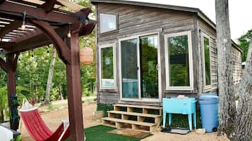 A rustic cabin with large windows, a wooden deck, a hammock, and a trash bin.