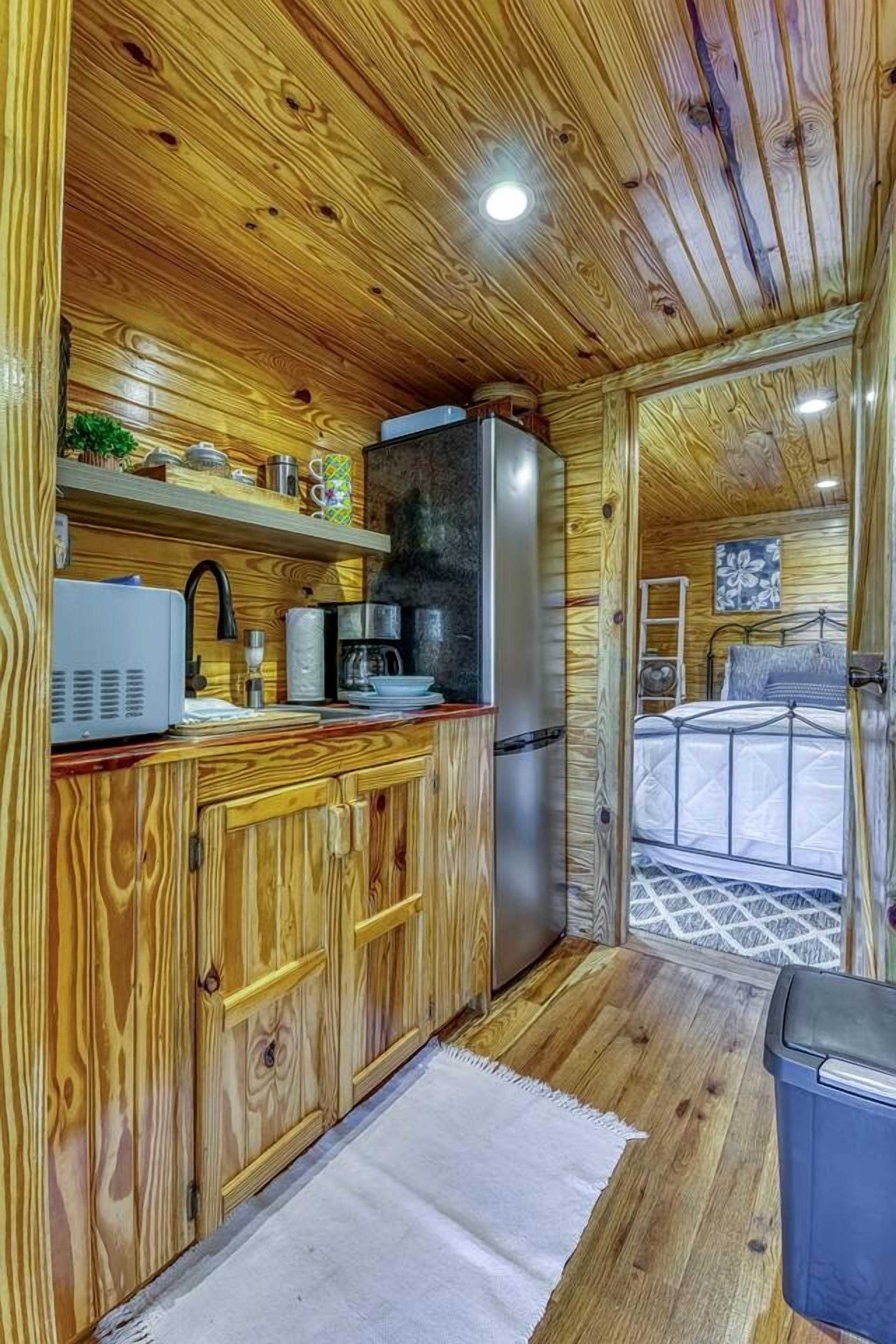 A wooden cabin interior with a kitchenette, a bed, and a view of the outside.