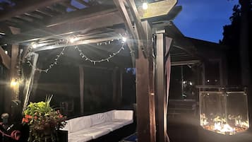 Een terras buiten met een houten pergola, sfeerverlichting, een vuurplaats en een zithoek.
