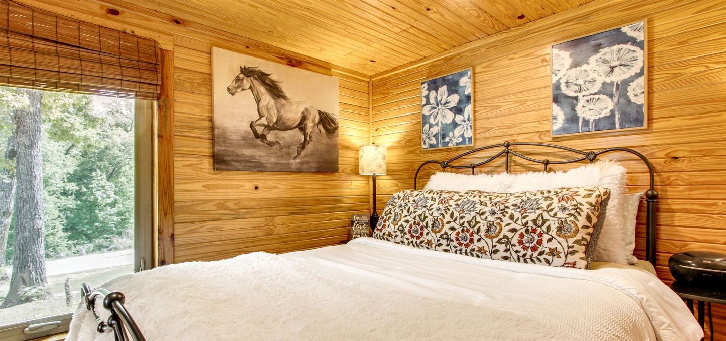 A wooden-paneled room with a bed, a painting of a horse, and two floral paintings on the wall.