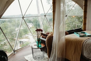 A bedroom with a large window, a bed, a small round table with a vase, and a view of greenery outside.