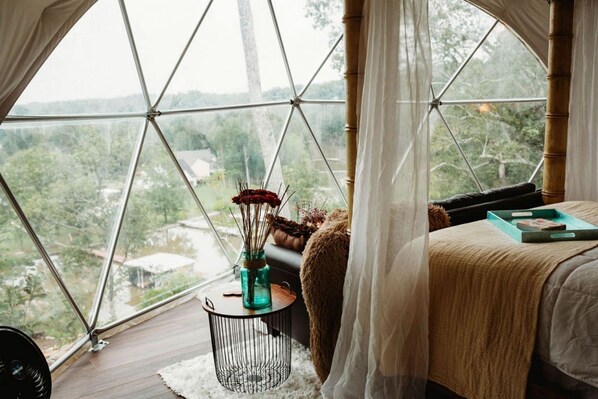 A bedroom with a large window, a bed, a small round table with a vase, and a view of greenery outside. - Gorgeous Tiny House with Indoor and Outdoor Lounging Space in Alabama (Rogersville)