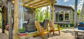 Una terraza de madera con dos sillas Adirondack, una parrilla y una cabaña pequeña con ventanas grandes.
