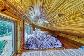 A bedroom with a bed, wooden walls, and a view through a large window.