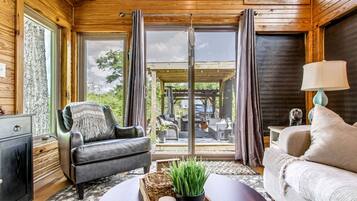 A cozy room with a leather armchair, a sofa, a television, and a view of the outdoors through large windows.