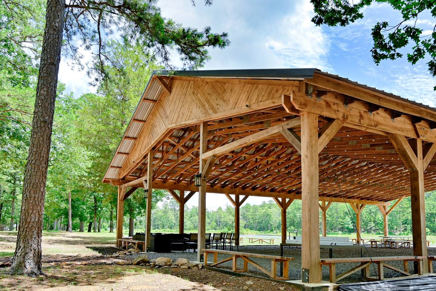 A wooden pavilion with a sloped roof, built on a gravel base, surrounded by trees.