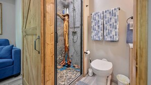 Um banheiro com box de vidro, vaso sanitário e um sofá azul na sala adjacente.