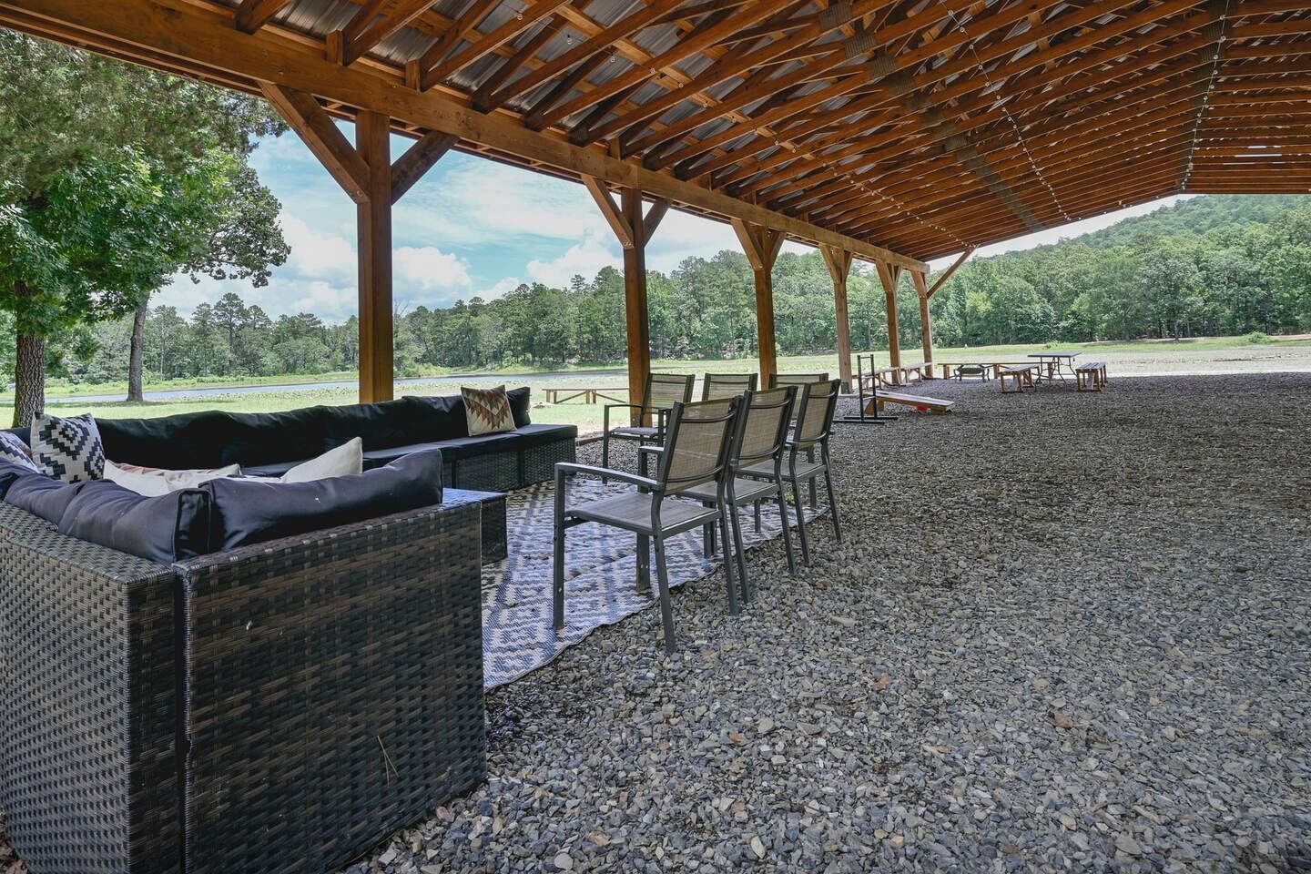 An outdoor covered seating area with wicker furniture and tables, gravel flooring, and a view of a green landscape.
