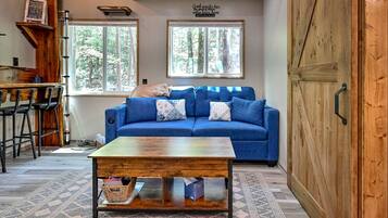 A cozy living room with a blue sofa, a wooden coffee table, and a view of trees through large windows.