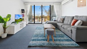 A modern living room with a grey sofa, a small wooden table, a blue and white rug, a potted plant, and a television on a white stand. - Executive and Spacious Apartment With Balcony (Melbourne)