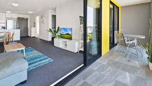 A modern living room with a flat-screen TV, a sofa, a dining table, and a kitchen in the background. - Executive and Spacious Apartment With Balcony (Melbourne)