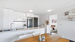 A modern kitchen with white cabinetry, a built-in island, and a dining area with a wooden table. - Executive and Spacious Apartment With Balcony (Melbourne)
