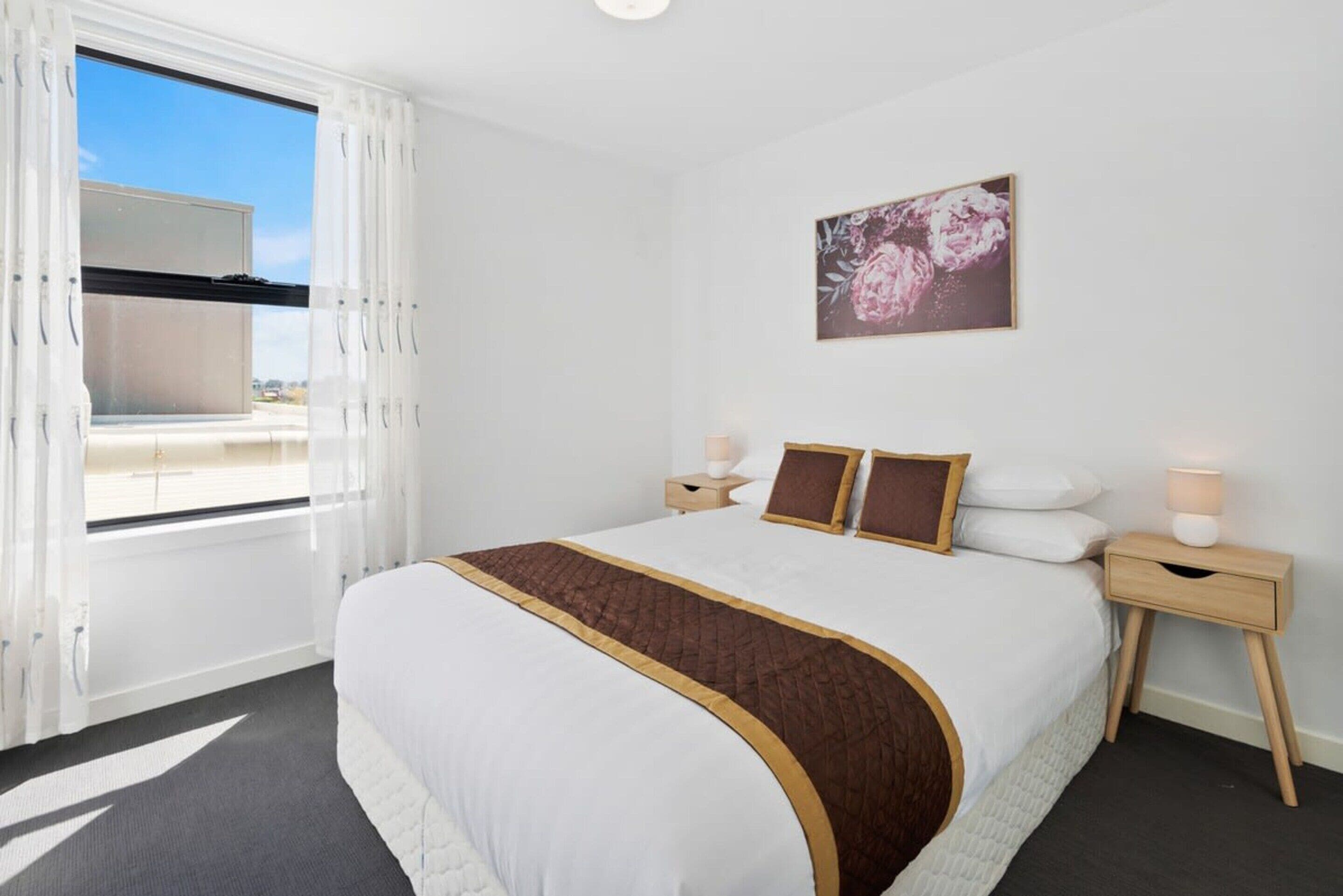 A hotel room with a large bed, a window with curtains, a bedside table with a lamp, and a floral artwork on the wall.