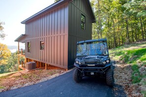 A two-story building with a covered outdoor area, a parked off-road vehicle, and a forested hillside.