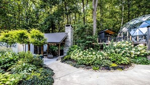 Ein modernes Haus mit einem Steinsschornstein, einer überdachten Terrasse und einem Garten mit verschiedenen Pflanzen.