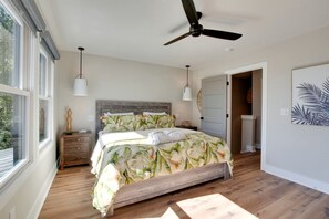 Un dormitorio con cama, piso de madera, ventilador de techo, lámparas de noche y una puerta que da a otra habitación.