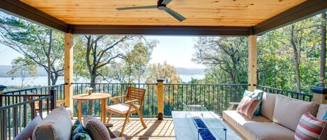 A covered outdoor seating area with a table, chairs, and a sofa, overlooking a body of water and trees.