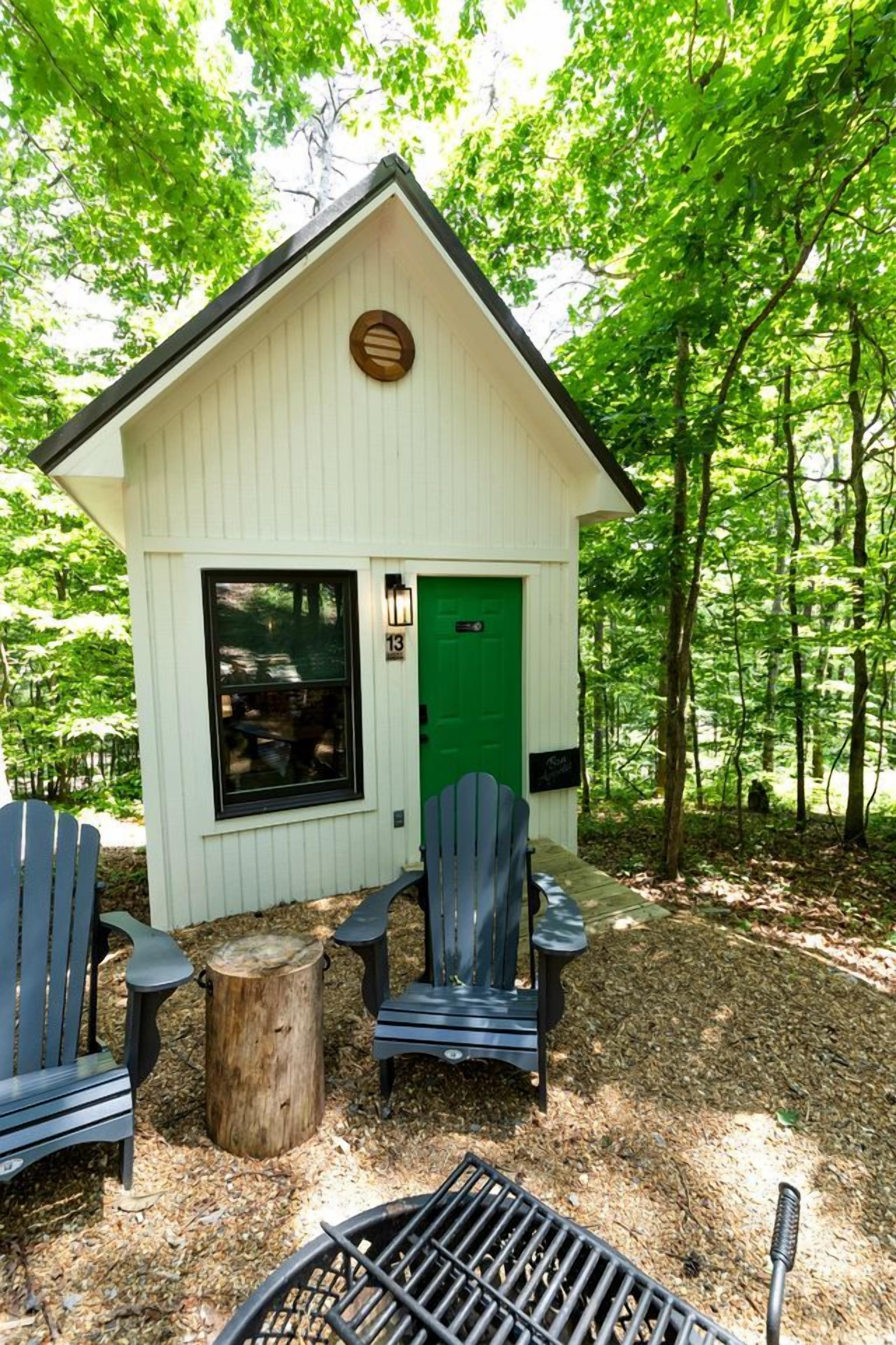 A small cabin with a green door, two Adirondack chairs, and a grill in front.