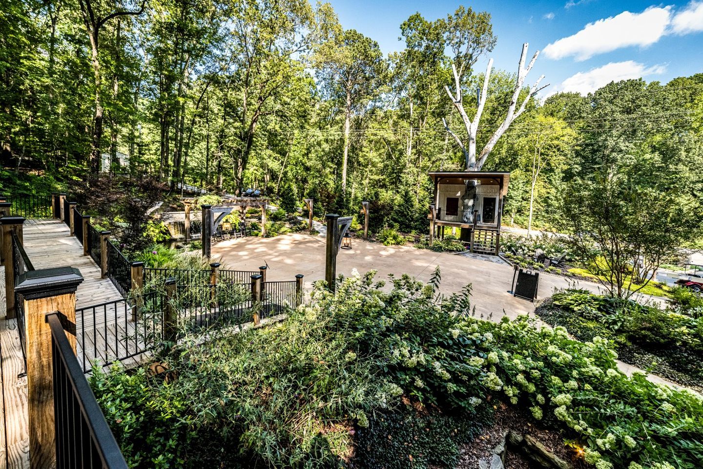 A wooden deck with a view of a forested area, a gazebo, and a paved area with a statue.