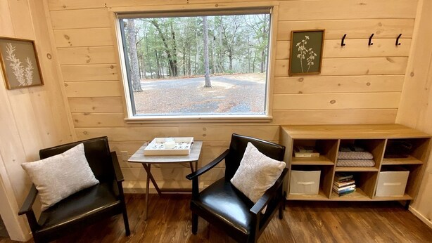 Una habitación acogedora con paredes de madera, una ventana con vistas a árboles, dos sillones con cojines, una mesita y una estantería.
