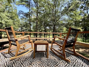 A wooden deck with three chairs and a small table, surrounded by patterned flooring and trees.