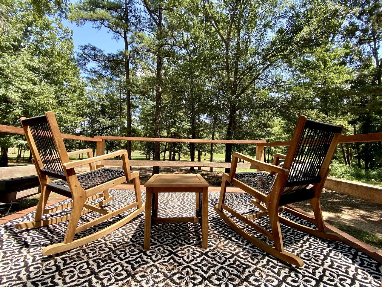 A wooden deck with three chairs and a small table, surrounded by patterned flooring and trees.