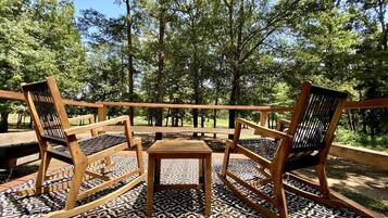 A wooden deck with three chairs and a small table, surrounded by patterned flooring and trees.