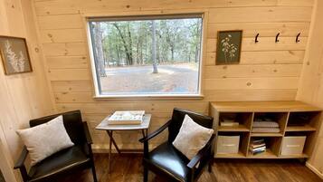 A cozy room with wooden walls, a window with a view of trees, two chairs with cushions, a small table, and a bookshelf.