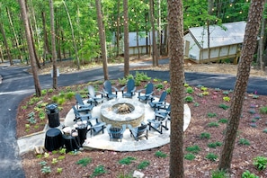 A circular outdoor seating area with chairs and a fire pit, surrounded by trees and a building with a metal roof.