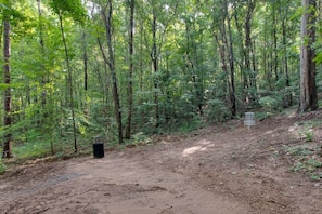 Zona boscosa con un sendero de tierra, un bote de basura metálico y un letrero.