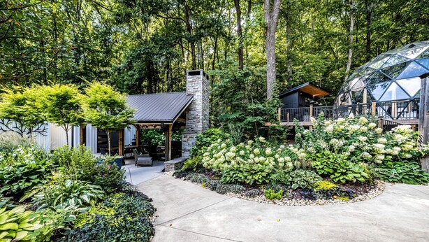 A modern house with a stone chimney, a covered patio, and a garden with various plants. - Off-grid Tree House Suitable for Four in Alabama (Scottsboro)