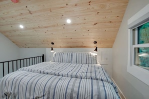 A bedroom with a sloped ceiling, a bed with striped bedding, two wall-mounted lights, and a window with a view.