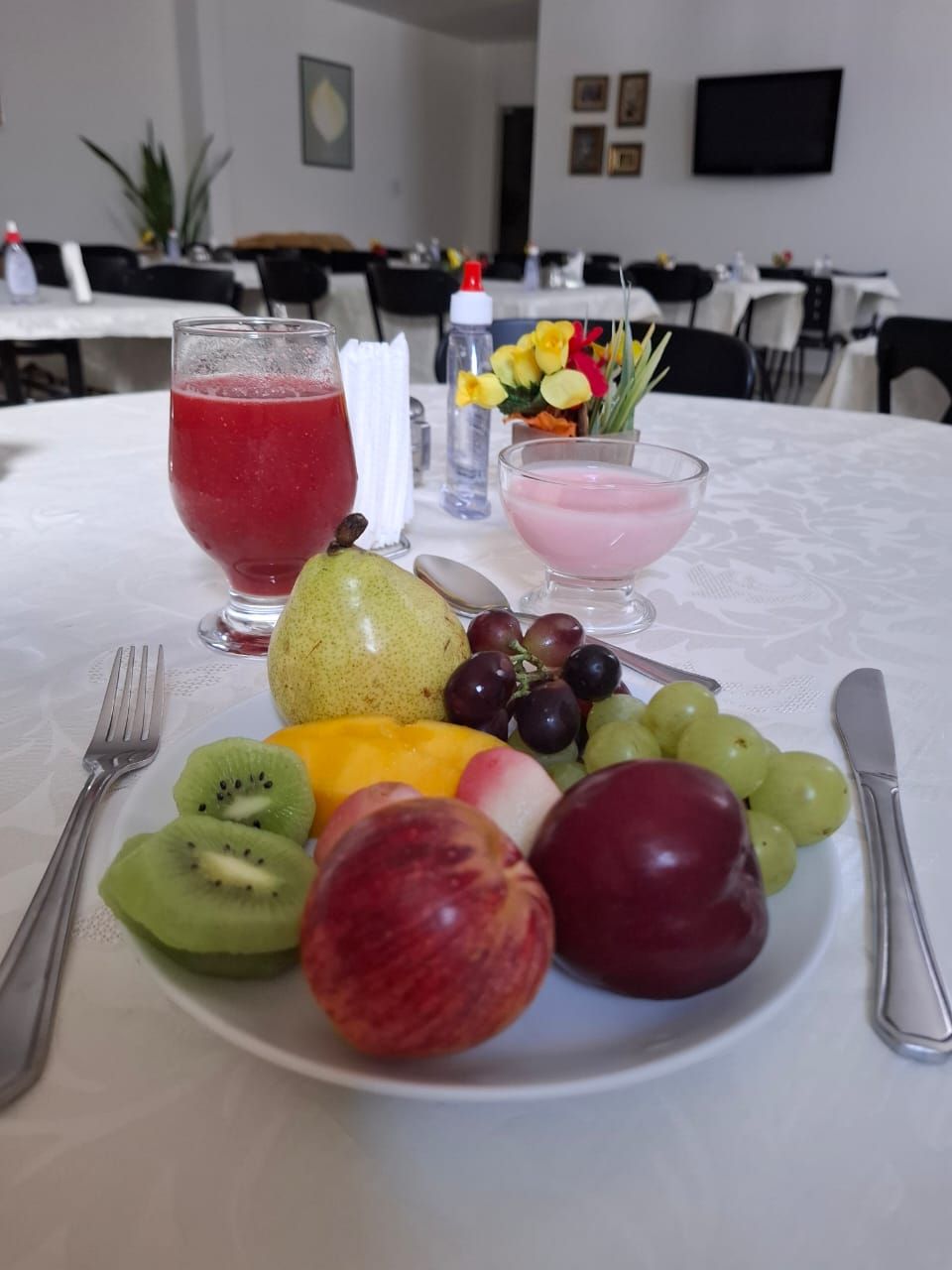 Une table avec une assiette de fruits, un verre de jus rouge, un verre d’une boisson rose et un petit vase de fleurs.