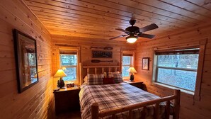 Acogedora habitación tipo cabaña con una cama, dos mesitas de noche con lámparas, un ventilador de techo y una ventana con vistas a árboles.
