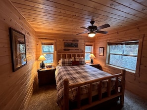 A cozy cabin bedroom with a bed, two bedside lamps, a ceiling fan, and a window with a view of trees. - Rustic Family Cabin Nestled Among the Towering Trees in Idaho (Island Park)