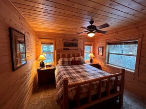Acogedora habitación tipo cabaña con una cama, dos mesitas de noche con lámparas, un ventilador de techo y una ventana con vistas a árboles.
