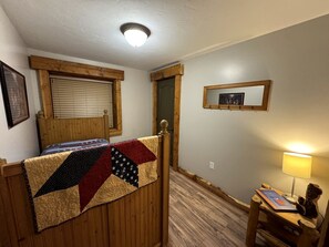 A room with a wooden bed, a window with blinds, a desk with a lamp, and a mirror on the wall.