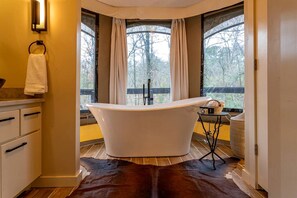 A modern bathroom with a freestanding bathtub, a wooden floor, and a view of trees through large windows.