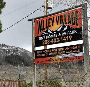Une enseigne pour Valley Village Tiny Homes & RV Park, avec les coordonnées et les services proposés, tels que des mini-maisons à louer ou à vendre, des tentes de luxe pour le « glamping », et des steaks à faire cuire au feu de camp.