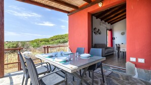 An outdoor dining area with a table set for four, surrounded by wicker chairs, a stone floor, and a red wall. - 'Villetta Wild' Apartment with Sea View, Shared Terrace and Air Conditioning (Stintino)