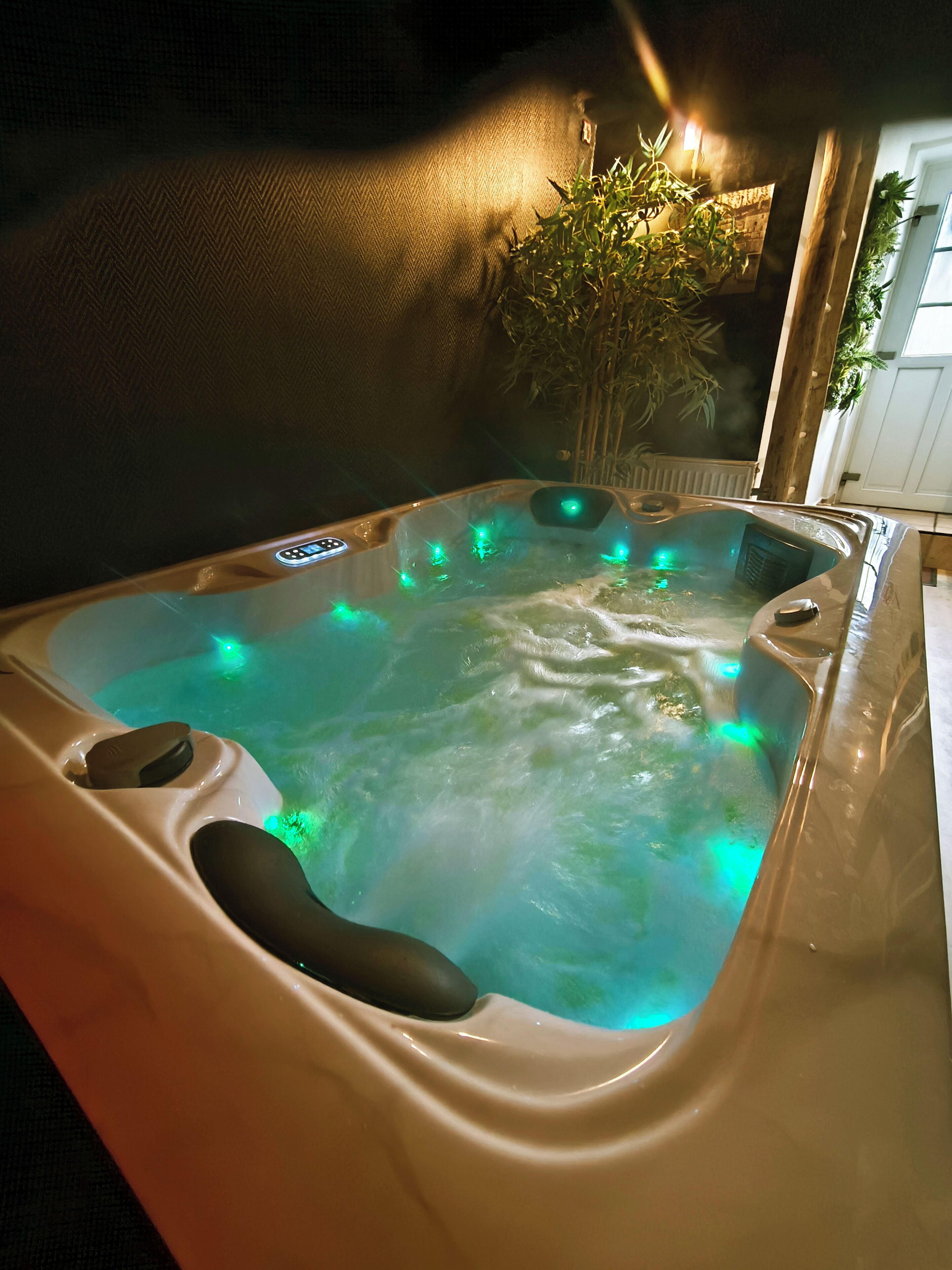 A hot tub with water jets and green lighting, surrounded by a dark wall and a plant.