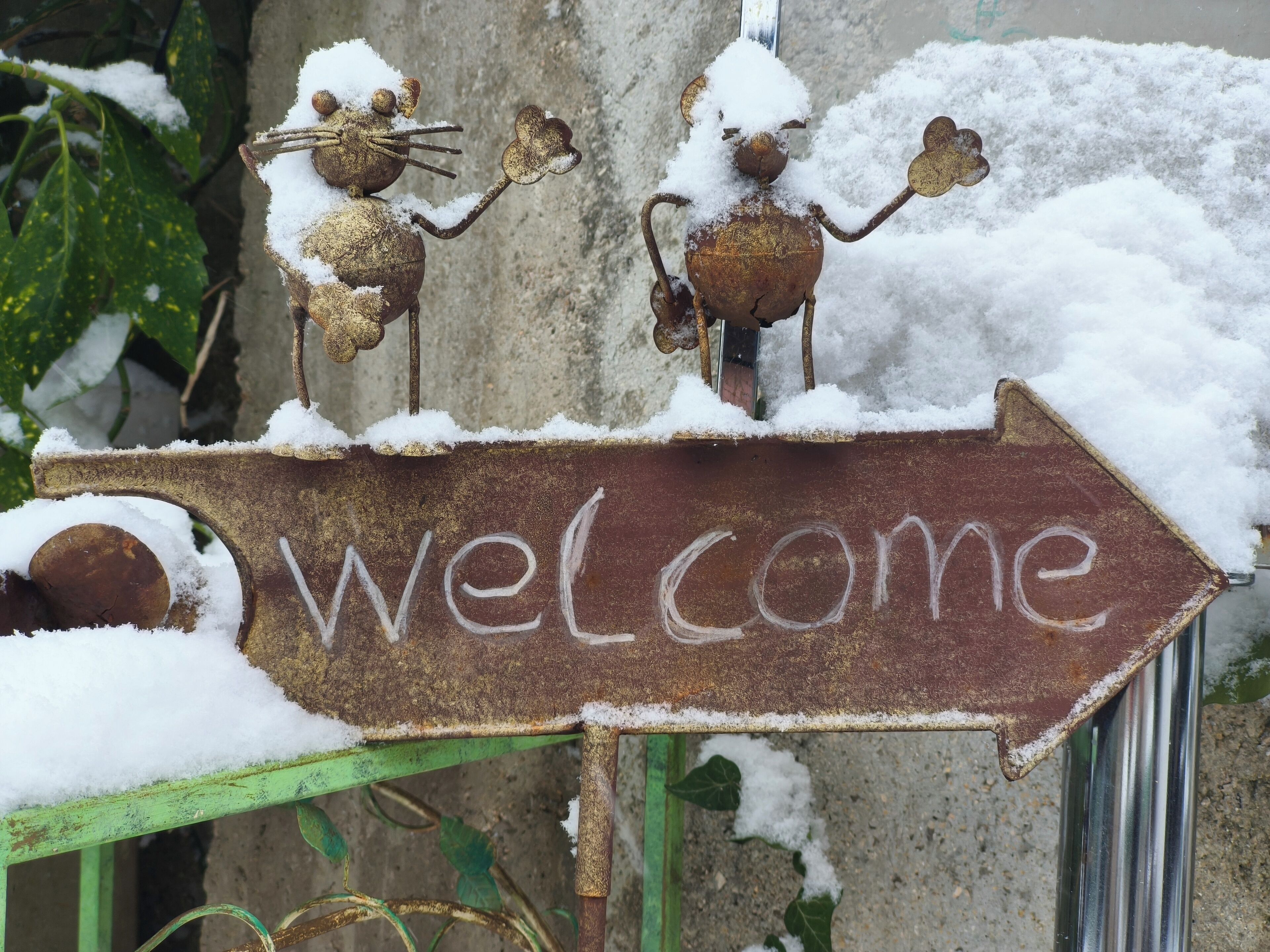 Une vieille enseigne métallique rouillée, surmontée d’une flèche indiquant « Bienvenue » et recouverte de neige, le tout entouré de neige et de verdure.