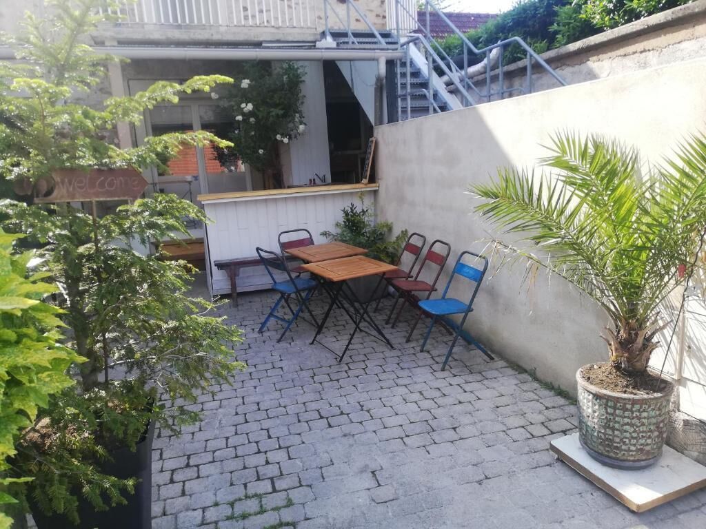 Een terras buiten met een houten tafel en blauwe stoelen, omgeven door potplanten en een betonnen muur.