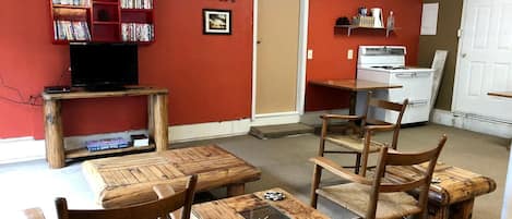 Una habitación con una pared roja, una estantería, un televisor, una mesa de comedor de madera con sillas y una zona de cocina con estufa.