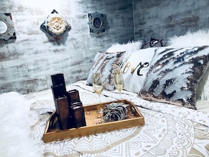 Una cama con una colcha estampada, una bandeja con botellas y vasos, y almohadones decorativos.