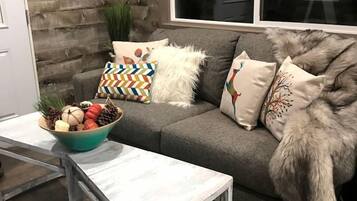 A living room with a grey sofa, white and patterned cushions, a white table, and a bowl of decorative items.