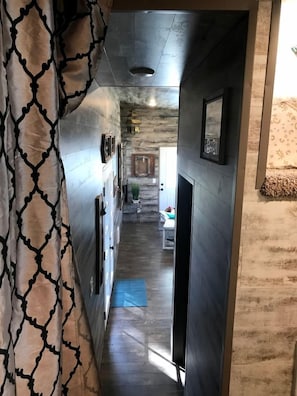 Un couloir étroit, orné d’un rideau à motifs, avec un mur en bois et une porte donnant sur une pièce où se trouve un lit.