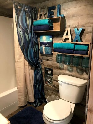 Ein Badezimmer mit Duschvorhang, Toilette, Ablagefläche mit gefalteten Handtüchern, einem gerahmten Bild und Holzbuchstaben, die 'REELAX' schreiben.