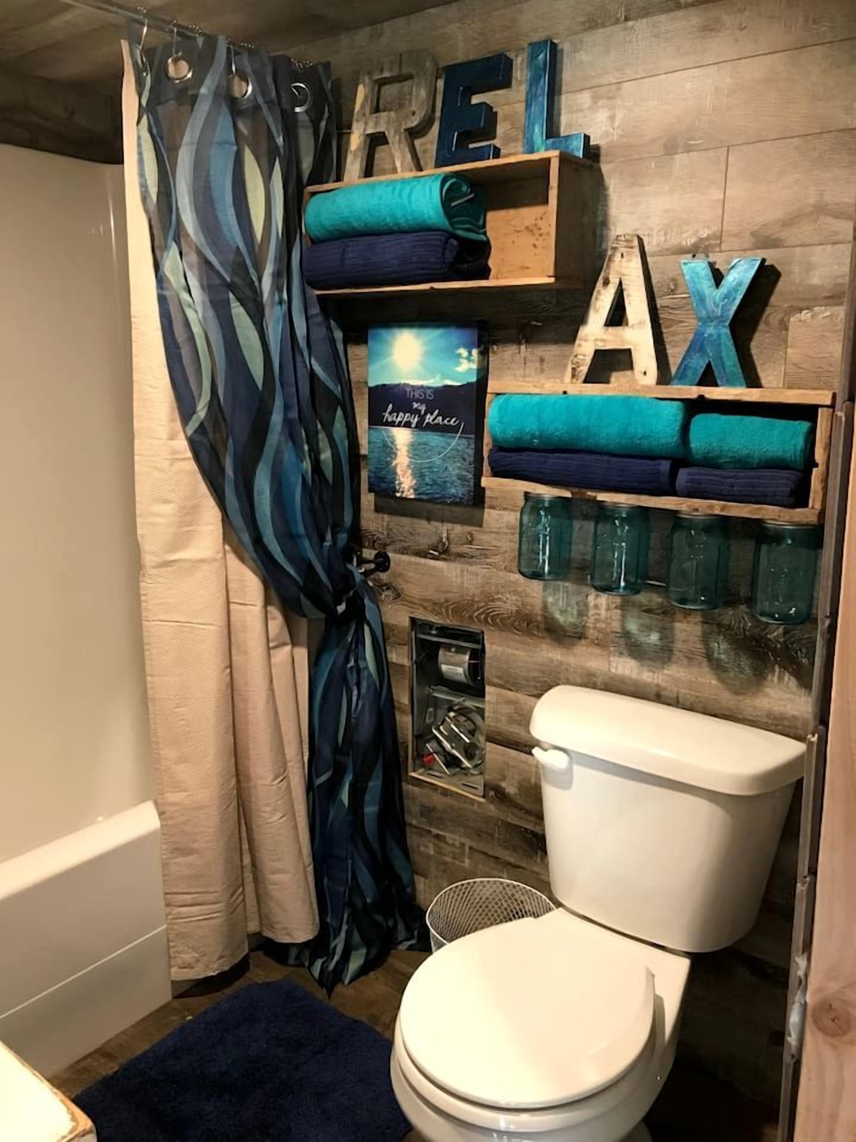 A bathroom with a shower curtain, a toilet, a shelf with folded towels, a framed picture, and wooden letters spelling 'REELAX'.