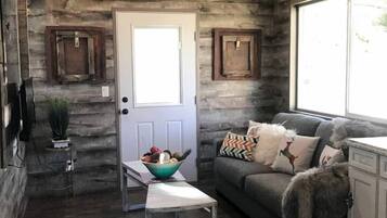 A rustic interior with a wooden ceiling, a white door, a grey sofa with patterned cushions, a small table with a bowl, and a window with a view.